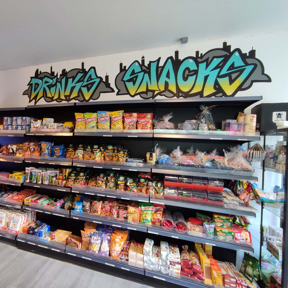Graffiti-Schriftzug 'Drinks' und 'Snacks' über einem Regal voller Snacks und Getränke in einem Kiosk in Bremerhaven."