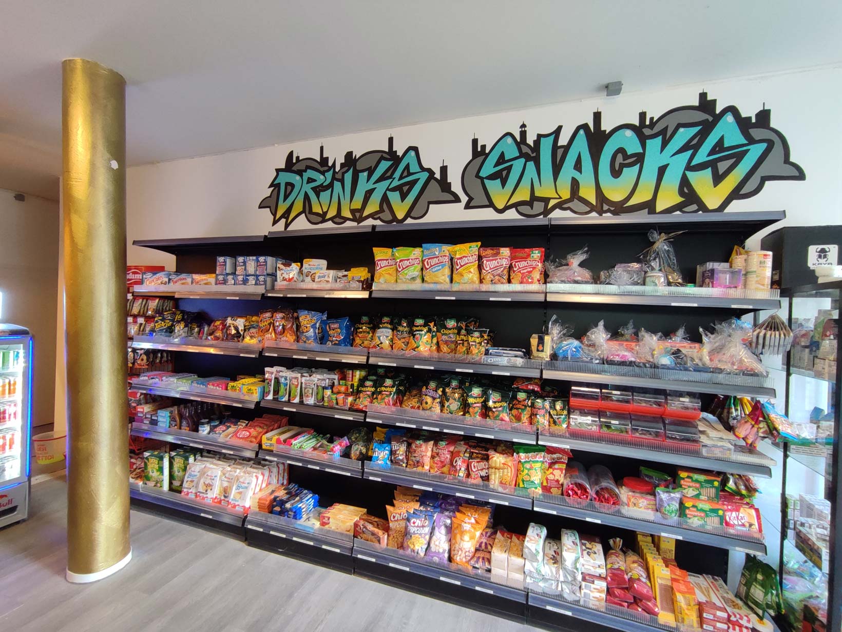 Graffiti-Schriftzug 'Drinks' und 'Snacks' über einem Regal voller Snacks und Getränke in einem Kiosk in Bremerhaven."