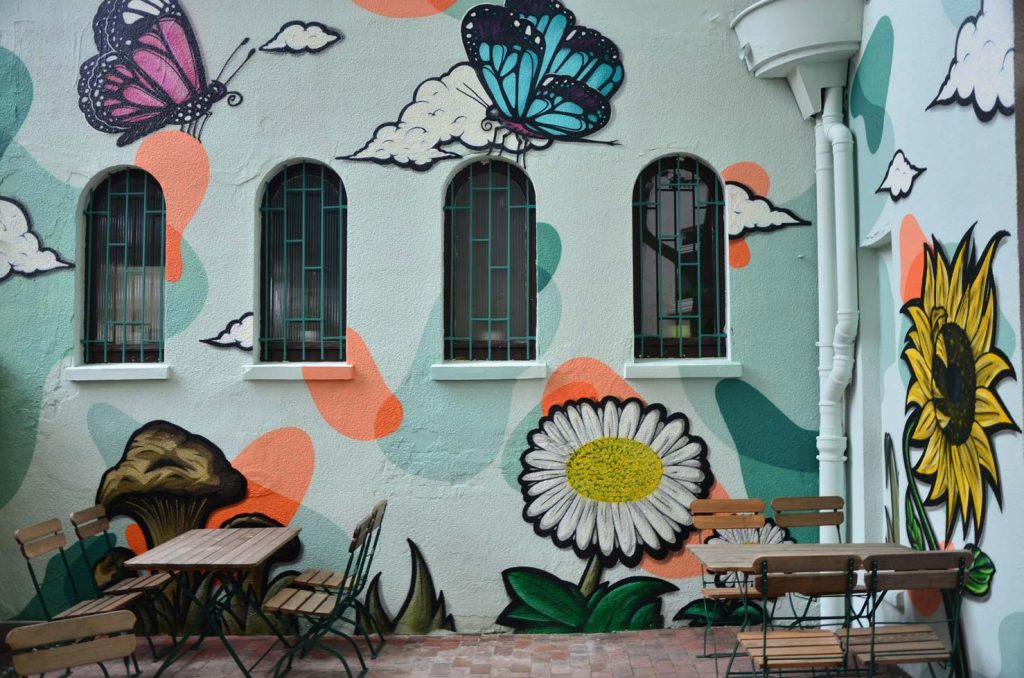 Auftrags-Graffiti für das Bio-Restaurant Seidenspinner in Oldenburg mit Schmetterlingen, einer Sonnenblume, Pilzen und Wolken im Hintergrund, umgeben von grafischen Formen.