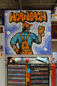 Ein farbenfrohes Graffiti im Hornbach-Markt zeigt einen Handwerker mit Latzhose, Sprühdose und Werkzeugen. Im Hintergrund sind Rahmenfertigungsartikel und Kunstbilder ausgestellt.