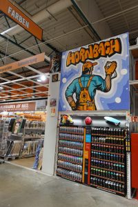 Ein farbenfrohes Graffiti im Hornbach-Markt zeigt einen Handwerker mit Latzhose, Sprühdose und Werkzeugen. Im Hintergrund sind Rahmenfertigungsartikel und Kunstbilder ausgestellt.