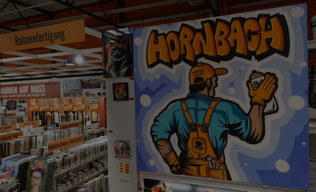 Ein farbenfrohes Graffiti im Hornbach-Markt zeigt einen Handwerker mit Latzhose, Sprühdose und Werkzeugen. Im Hintergrund sind Rahmenfertigungsartikel und Kunstbilder ausgestellt.